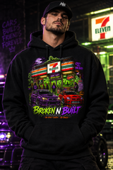 A stylized Halloween-themed storefront scene featuring a 7-Eleven sign and building facade, with purple and red cars in the foreground, glowing green ghosts, and large text reading 'BROKEN N BUILT' and 'We Don't Sleep.